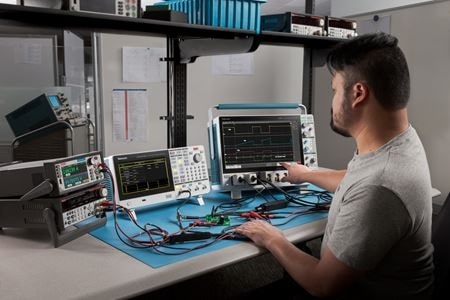 A technician at a workbench using an oscilloscope, function/waveform generator, digital multimeter, source measure unit, and power supply.