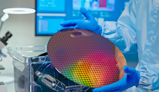 Technician in cleanroom attire holding a semiconductor wafer with colorful reflections, in a lab with a microscope